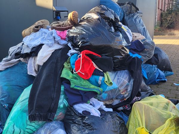 Vor zwei großen, grauen Altkleidercontainern steht ein großer Haufen aus verschiedenen Säcken und losen Kleidungsstücken. Die Säcke sind überwiegend schwarz, daneben sind auch bunte Kleidungsstücke sichtbar, darunter ein rotes und ein grünes Kleidungsstück.
