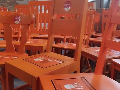 Mehrere orangefarbene Holzstühle stehen dicht beieinander in einem Raum mit großen Fenstern und einer Wand aus Naturstein. Auf den Sitzflächen der Stühle liegen quadratische orangefarbene Pappschilder mit schwarzem Rand und weißer Schrift. Die Rückenlehnen der Stühle tragen runde Aufkleber mit weißer Schrift auf rotem Grund. Im Hintergrund ist ein Holzgitter und ein Ventilator an der Wand zu sehen.