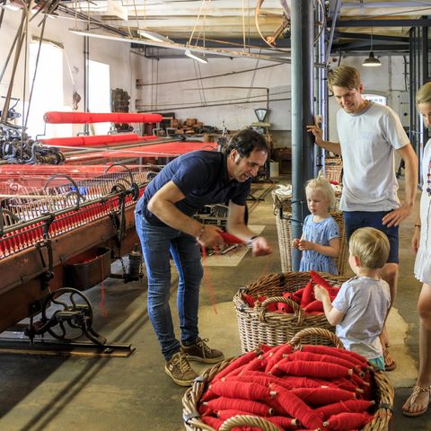 Eine Familie besucht einen historischen Webstuhl im Tuchmacher Museum Bramsche im Osnabrücker Land. Ein Guide erklärt den Kindern anschaulich, wie rote Stoffe hergestellt werden. Die alten Webstühle und das industrielle Ambiente lassen Geschichte lebendig werden und bieten ein spannendes Ausflugsziel für Groß und Klein.