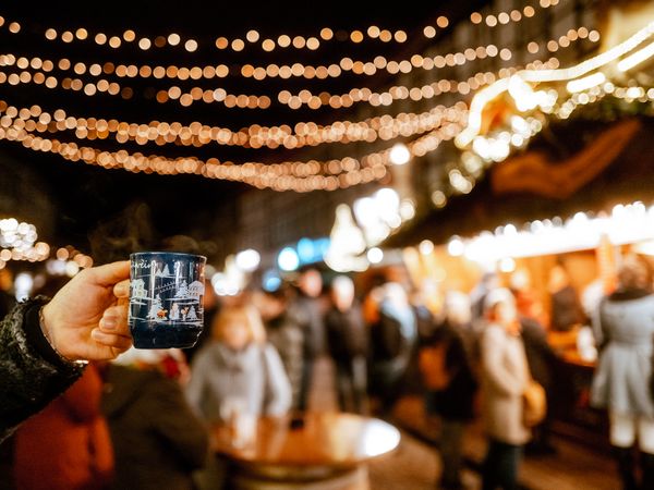 Weihnachtsdorf in Einbeck im Lichterglanz Hand, die eine Tasse Glühwein hält im Fokus und das beleuchtete Einbecker Weihnachtsdorf dahinter.