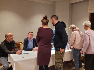 Samer Tannous und Gerd Hachmöller sitzen an einem Tisch mit weißer Tischdecke, vor ihnen stehen mehrere Personen in einem Raum mit Fenster und Pflanze.