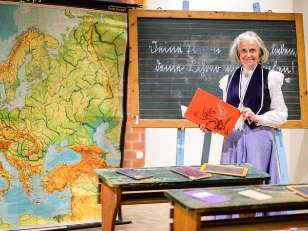 Eine Lehrerin steht in einem historischen Klassenraum in der Ausstellung "Von Federkiel und Rechenschieber"