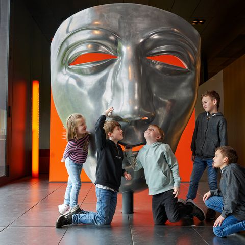 Kinder bestaunen eine große, silberne Varusmaske im Museum und Park Kalkriese im Osnabrücker Land. Die leuchtend orangefarbenen Augen der Maske und die moderne Ausstellung sorgen für ein spannendes und lehrreiches Erlebnis für die ganze Familie.