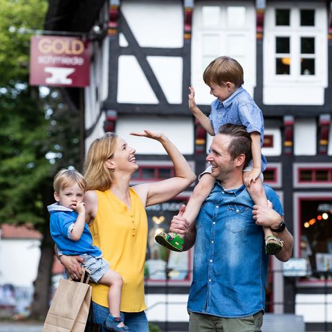 Eine fröhliche Familie steht vor einem Fachwerkhaus im Osnabrücker Land. Die Mutter und der Vater lachen, während der Vater den älteren Sohn auf den Schultern trägt und die jüngere Tochter von der Mutter gehalten wird. Das historische Gebäude im Hintergrund verleiht der Szene einen charmanten, regionalen Charakter.