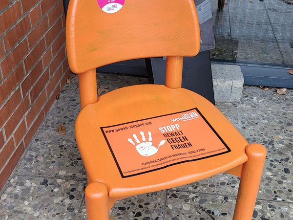 Oranger Stuhl steht vor dem Rathaus. Auf der Sitzfläche ein Aufkleber mit weißer Hand und Text 'STOPP GEWALT GEGEN FRAUEN'. Auf der Rückenlehne ein runder rosa Aufkleber mit der Nummer '116 016'.