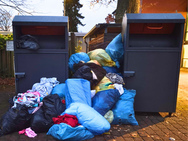 Zwei dunkelgraue Altkleidercontainer stehen auf einem gepflasterten Boden vor einem Baum. Vor den Containern liegen zahlreiche blaue, schwarze und gelbe Säcke sowie lose Kleidungsstücke, die den Bereich vor den Containern teilweise blockieren. 